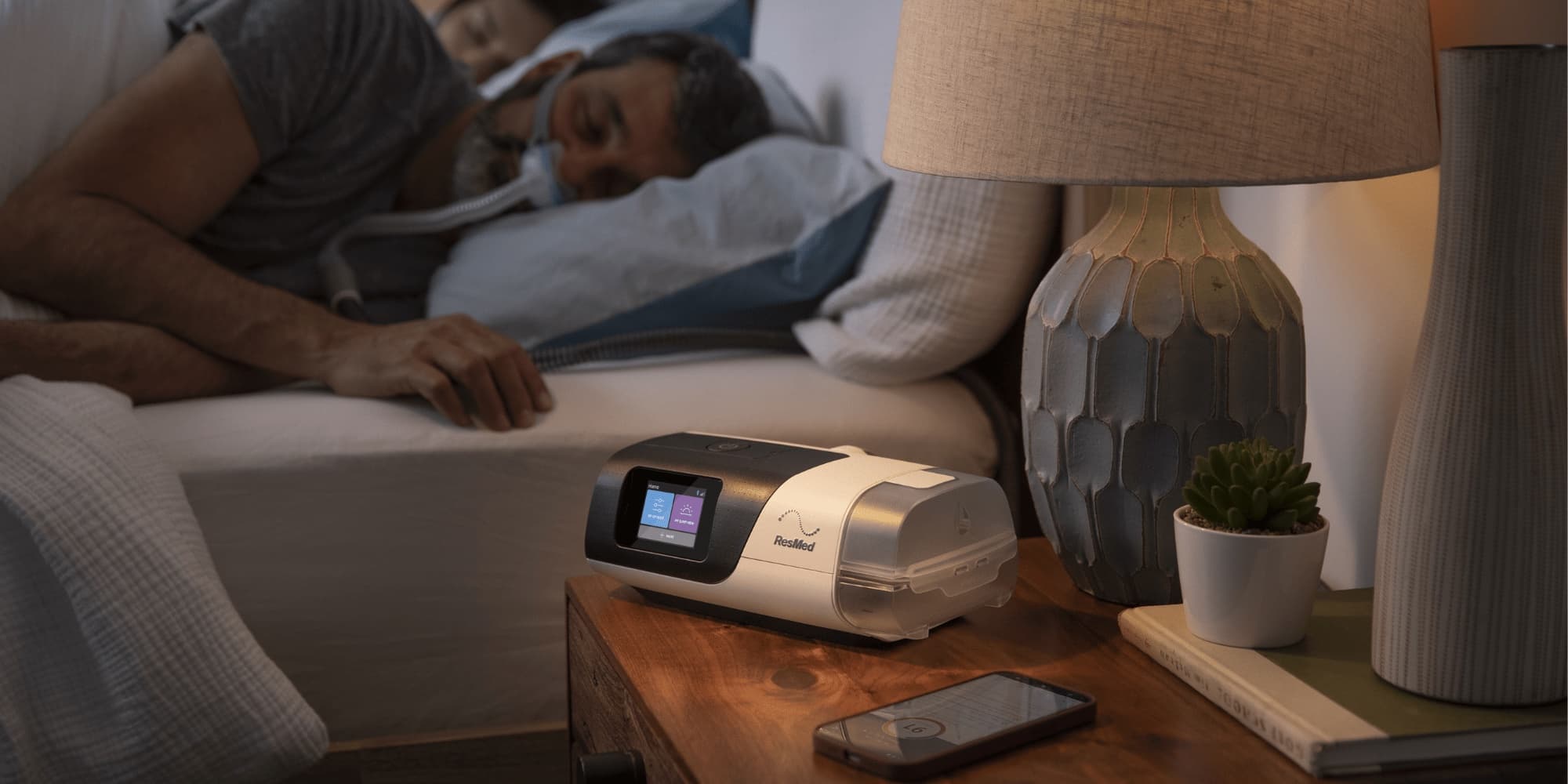 Aparat CPAP Resmed AirSense 11 na szafce nocnej, pacjent śpiący w masce